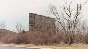 Two pictures of screentower surounded by overgrown brush.  Screen has peeling paint and faces an apartment complex.