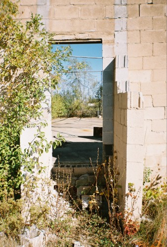 Overgrown and crumbling opening in a cinder block wall near the screen. Passage leads to the batting cages.