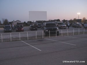 Cars are gathering in the gravel lot on a Saturday night in April.