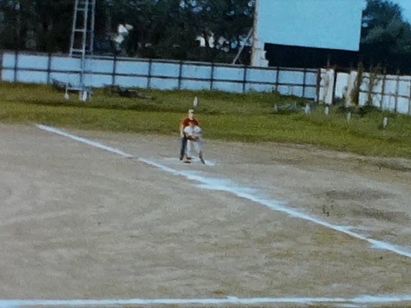 Still frame from 8mm home movie of a kids baseball game held on a field next to the El Rancho.  You can see the El Rancho screen in the background.
