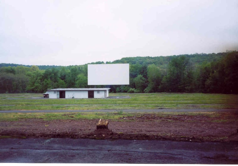 Field, screen and projection/concession building taken from the entrance road.  There used to be a natural tree barrier and short wooden fence blocking this view (note the tree stump in the foreground).