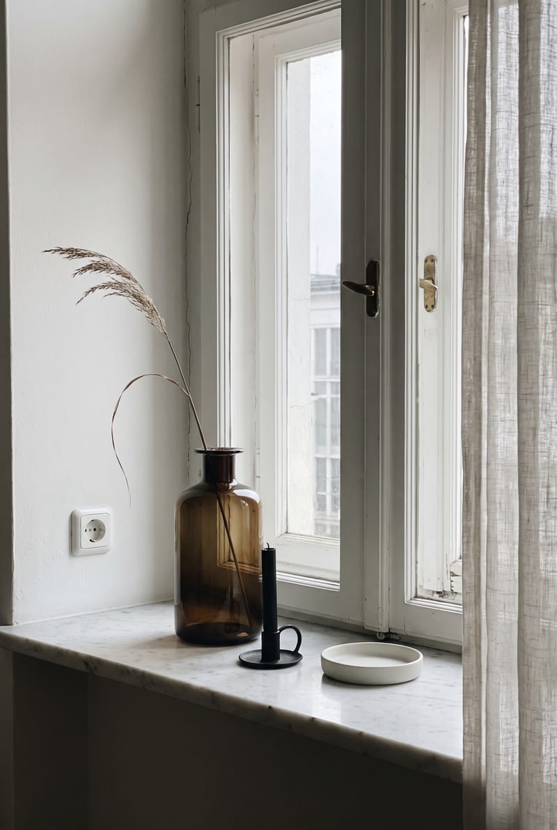 Fensterbank Deko Idee im modernen Stil: bernsteinfarbene Glasvase mit Trockengras, schwarze Kerze und flache Schale auf Marmor-Fensterbank