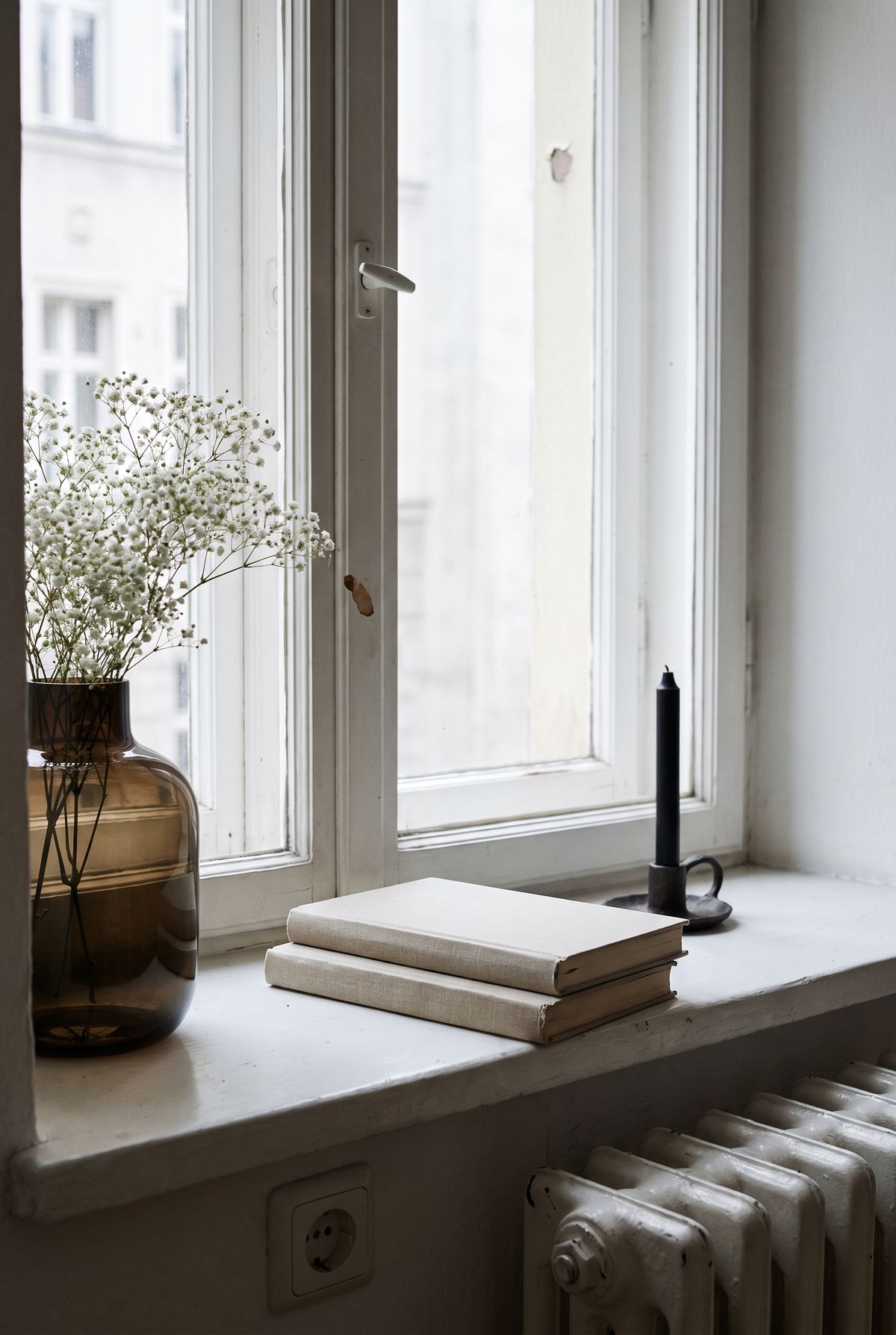 Fensterbank Deko Idee im Warm Minimal Stil: Rauchglasvase mit Schleierkraut, zwei liegende Bücher und schwarze Kerze auf weißer Altbau-Fensterbank