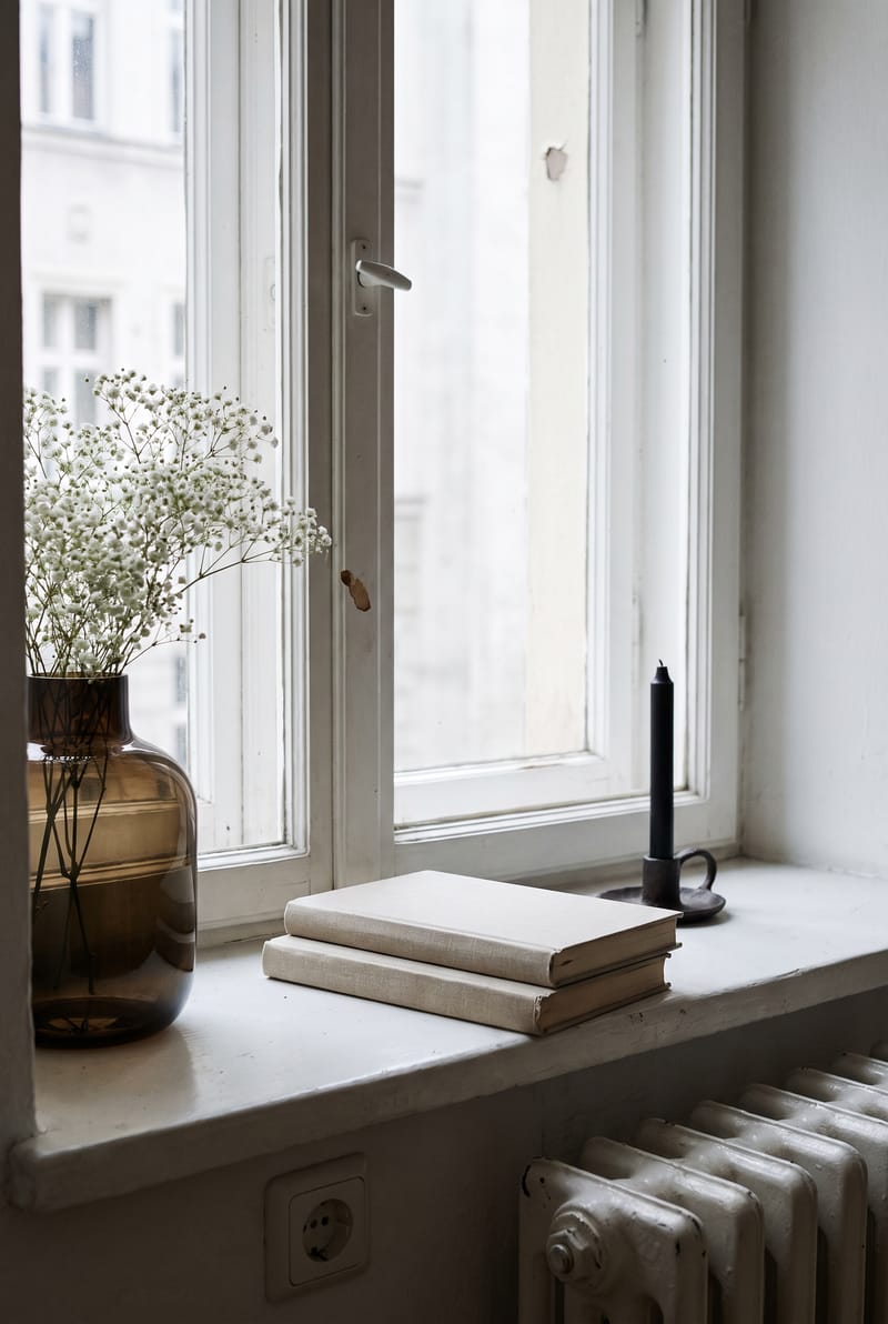 Fensterbank Deko Idee im Warm Minimal Stil: Rauchglasvase mit Schleierkraut, zwei liegende Bücher und schwarze Kerze auf weißer Altbau-Fensterbank