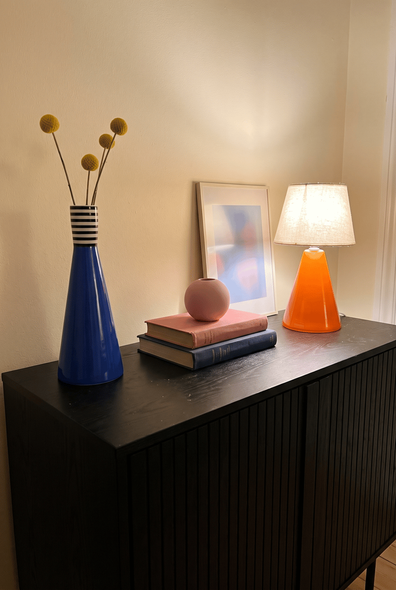 Sideboard Deko Idee im Dopamin Stil: kobaltblaue Kegelvase mit Craspedia, rosa Kugelvase auf Büchern und orange Glaslampe auf schwarzem Sideboard