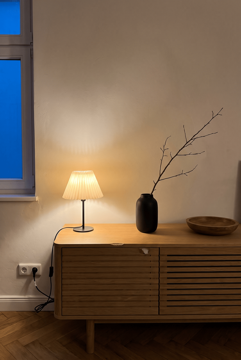 Sideboard Deko Idee im Japandi Stil: cremefarbene Faltenlampe, schwarze Vase mit Zweig und Holzschale auf Eichen-Sideboard am Abend