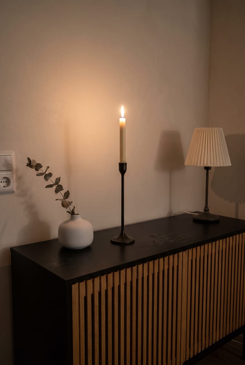 Sideboard Deko Idee im modernen Stil: hoher Kerzenständer mit brennender Kerze, weiße Vase mit Eukalyptus und kleine Lampe auf schwarzem Sideboard