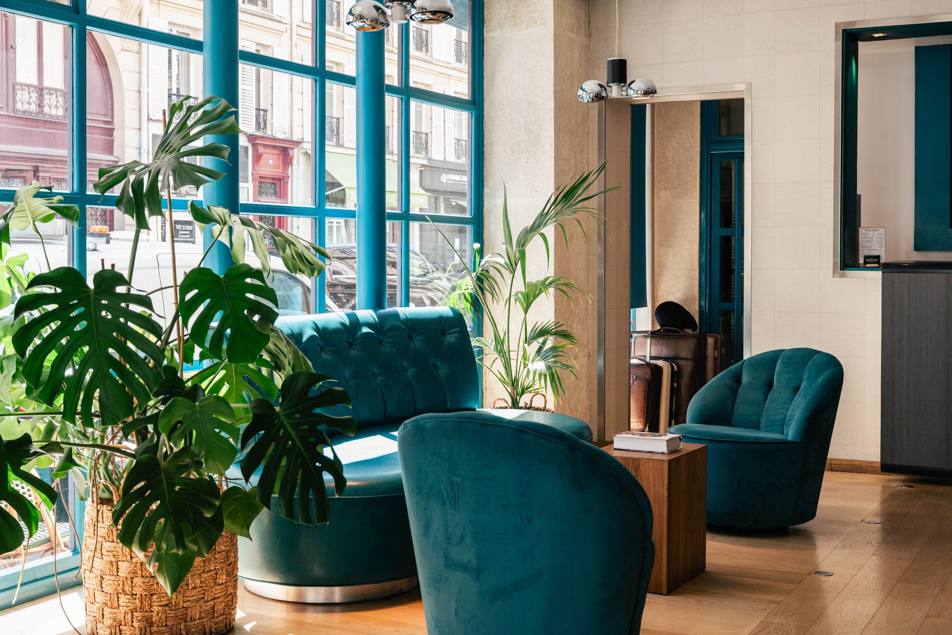 Salon de réception avec fauteuils en velours bleu, plantes vertes et baies vitrées donnant sur la rue.