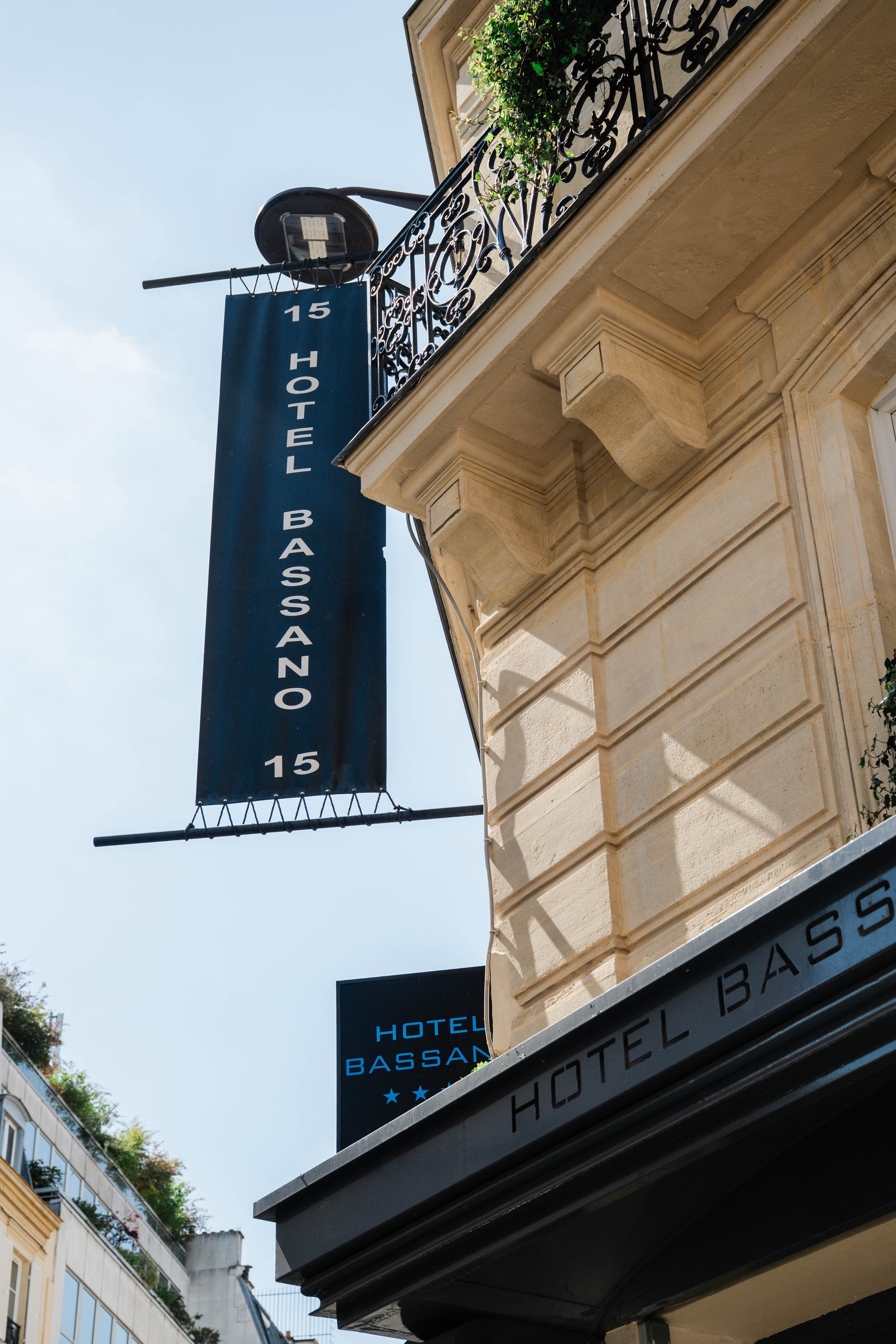 Enseigne verticale bleue de l'Hôtel Bassano sur une façade classique parisienne avec balcon.