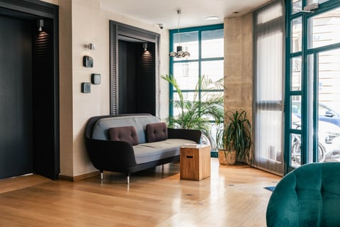 Salon d'hôtel avec canapé gris et noir, plantes vertes, porte-fenêtre donnant sur la rue, sol en bois.