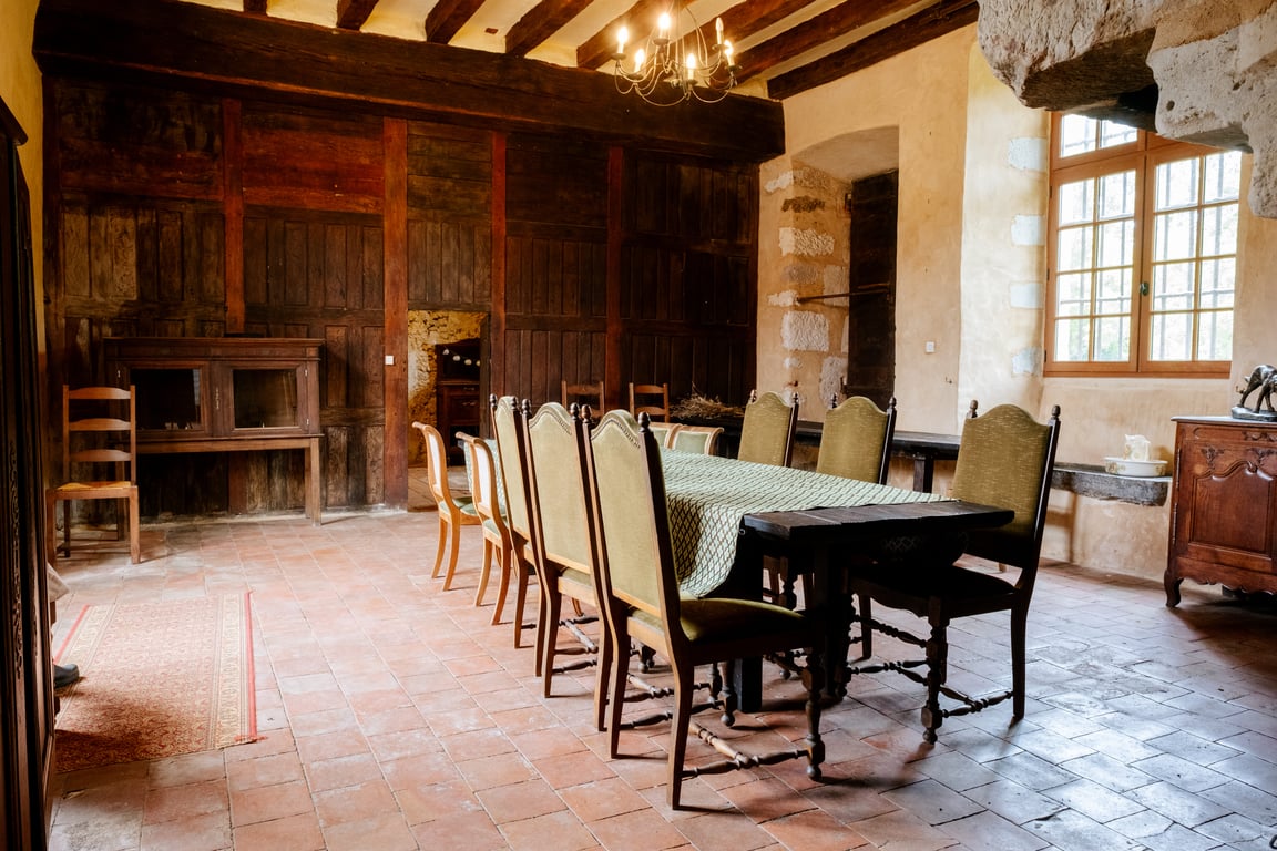 Salle de réception rustique avec poutres apparentes, grande table dressée, chaises en bois et mobilier ancien.