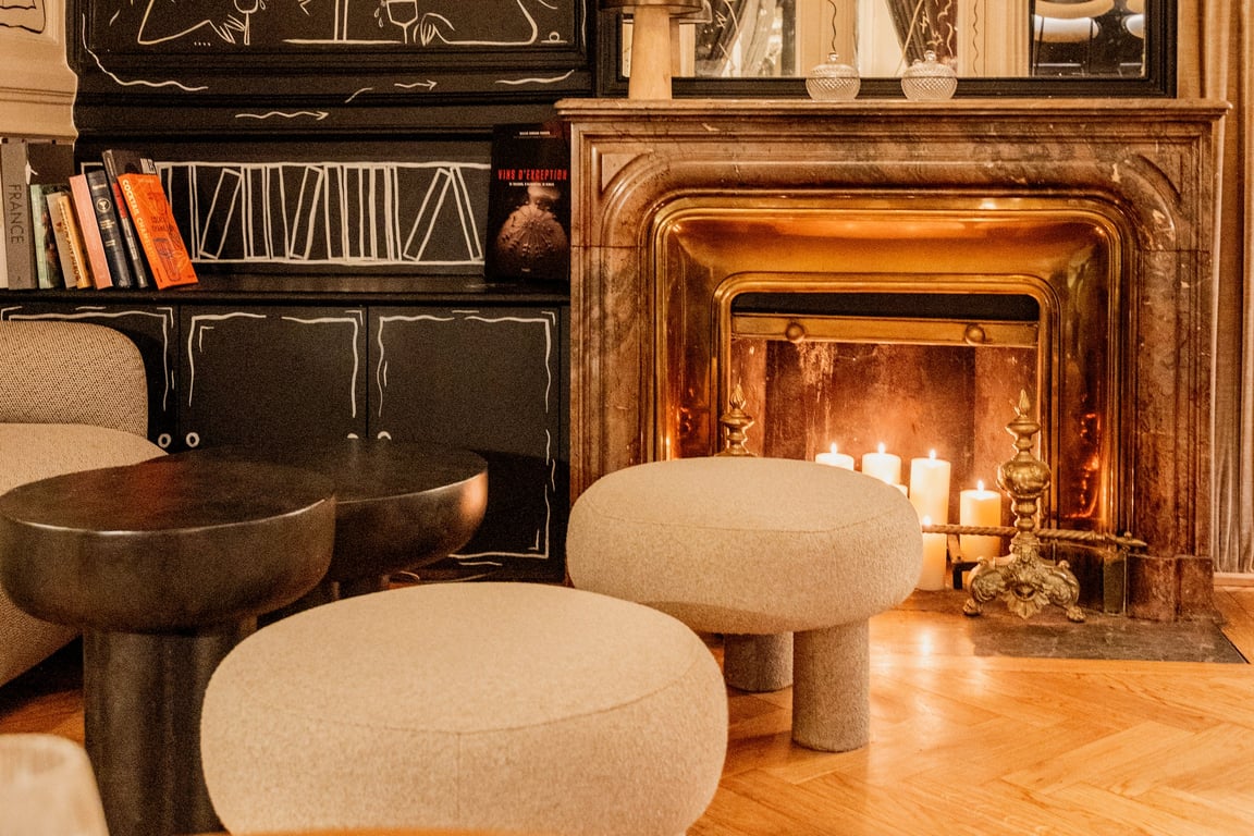 Salon luxueux avec cheminée dorée allumée, poufs beige, bibliothèque noire et décoration contemporaine.