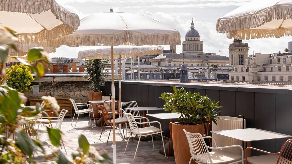 Rooftop terrace offering sweeping panoramic views of the city, furnished with beige parasols, contemporary furnishings, and framed by historic architecture beyond.