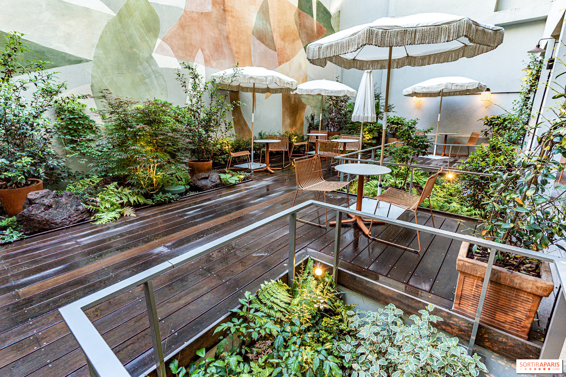 A sheltered courtyard featuring sunshades and wooden seating amongst verdant foliage, anchored by a vibrant abstract art wall.