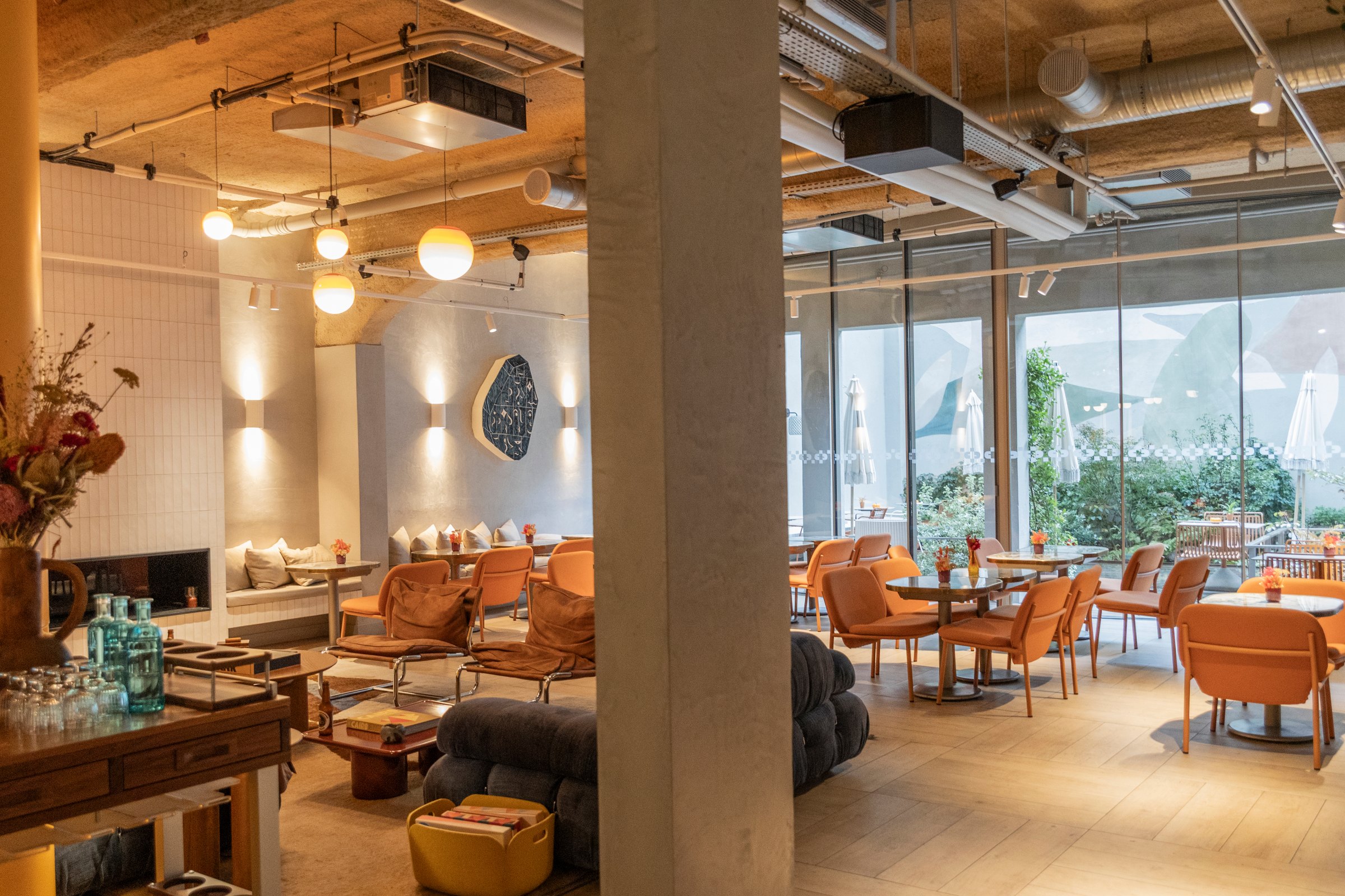 Contemporary lounge featuring vibrant orange seating, timber ceiling, suspended lighting fixtures, and sweeping mountain vistas framed by expansive floor-to-ceiling windows.