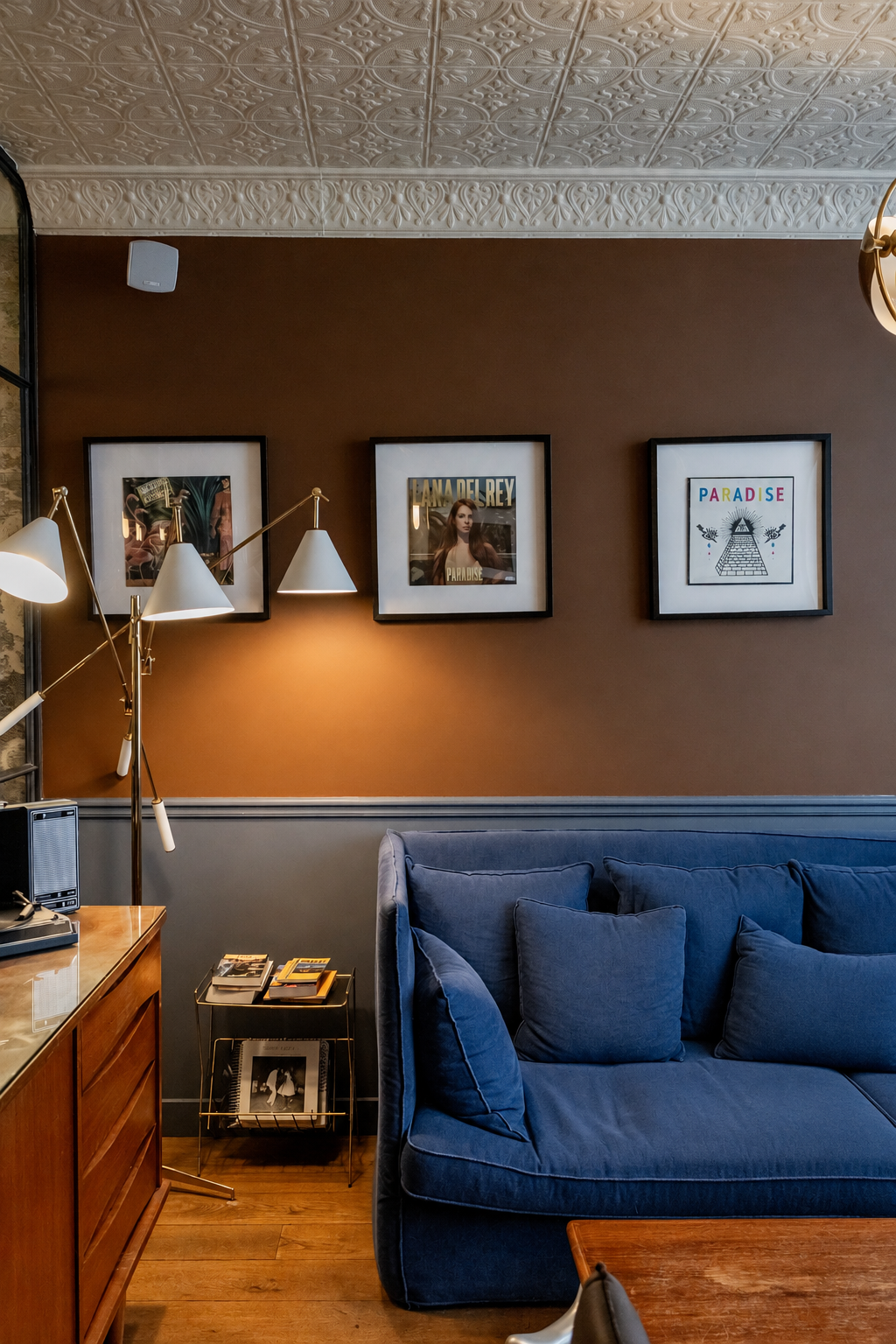 Hotel room featuring a navy blue sofa, soft taupe walls, three framed artworks displayed above the sofa, patterned white ceiling, and gilt floor lamp.