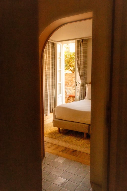 Chambre avec lit blanc visible à travers une porte cintrée, fenêtres ouvertes sur terrasse ensoleillée.