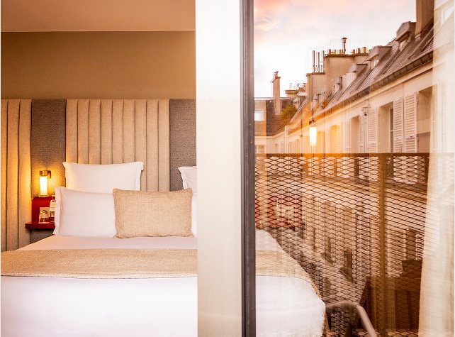 Chambre lumineuse ouverte sur un balcon et les toits de la ville au coucher du soleil