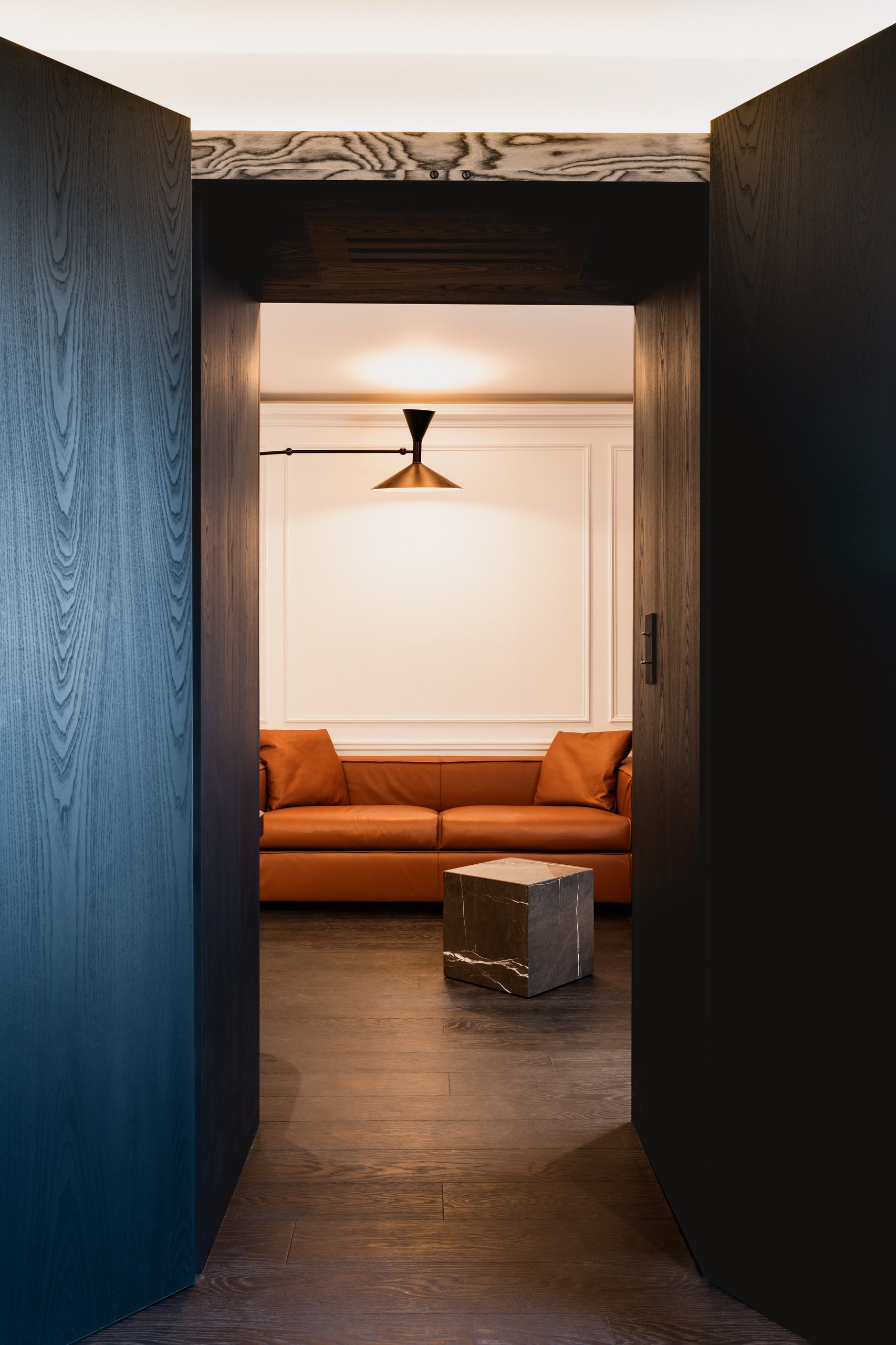 Hallway with open wooden doors revealing a lounge furnished with an orange sofa, small black table, and ceiling fan.