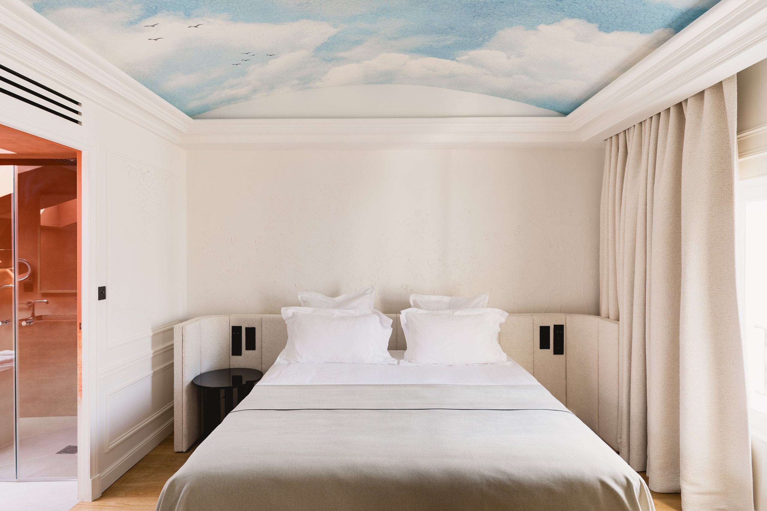 Chambre d'hôtel moderne avec lit double, plafond peint ciel bleu nuageux, salle de bain attenante et rideaux beiges.