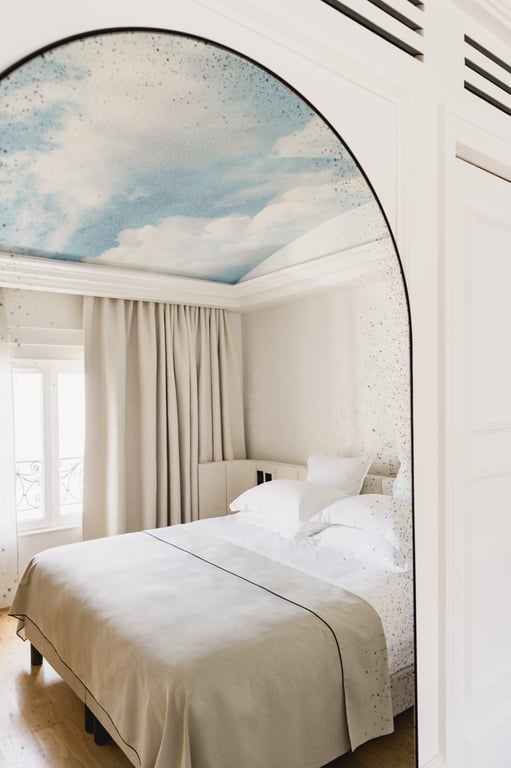 Chambre spacieuse avec lit double aux draps blancs, plafond peint ciel nuageux, rideaux beige et fenêtre donnant sur balcon.