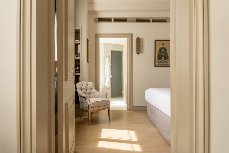 Chambre d'hôtel spacieuse avec lit blanc, fauteuil beige, salle de bain attenante et décoration classique épurée.
