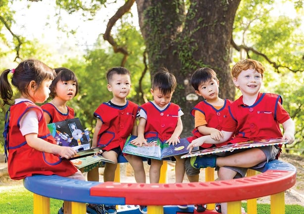 Young children sitting outdoors under a large tree, reading picture books together in a cheerful setting.