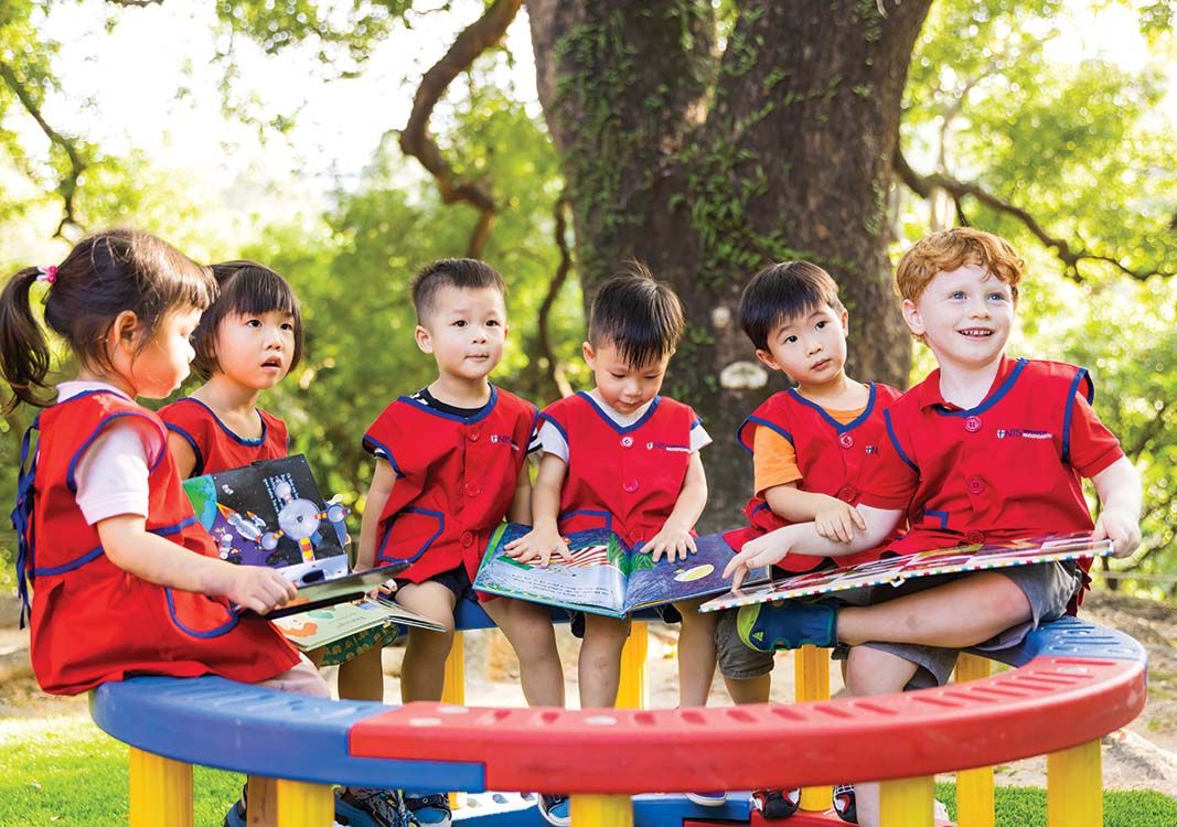 Young children sitting outdoors under a large tree, reading picture books together in a cheerful setting.