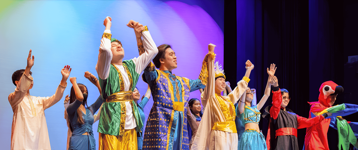 A group of students performs on stage in colourful costumes, holding hands and taking a bow after their show.