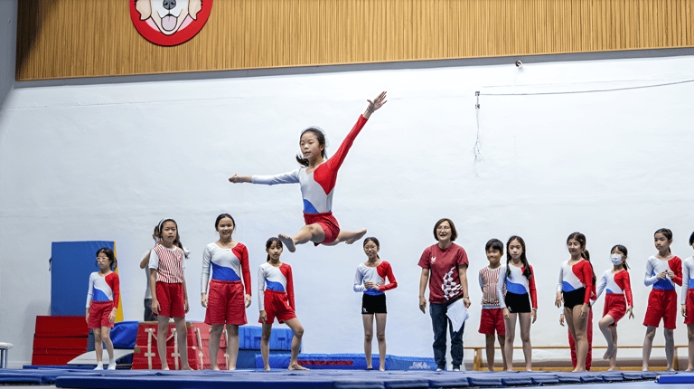 A student performs a leap during a gymnastics practice while classmates and teachers watch.