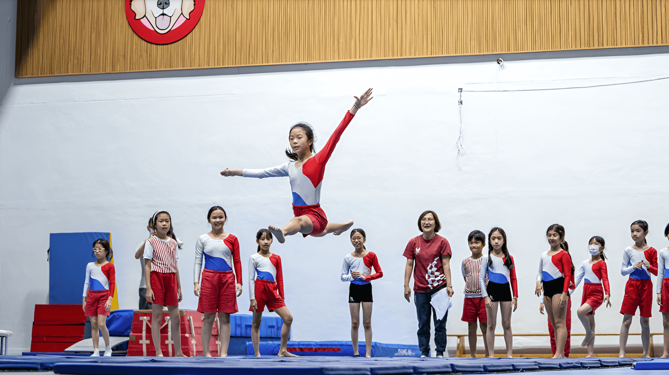 A student performs a leap during a gymnastics practice while classmates and teachers watch.
