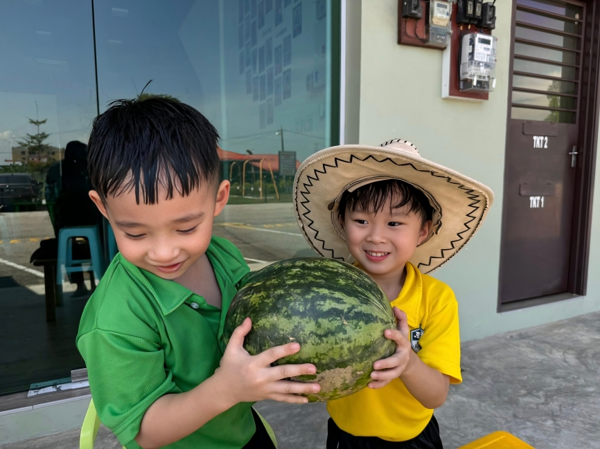Two young students smile as they explore the texture and weight of a watermelon, part of a fun outdoor learning experience.