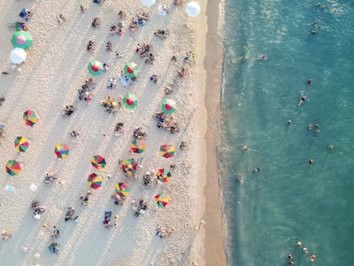 Imagem de uma praia vista de cima, com a água límpida, areia branquinha e várias pessoas.