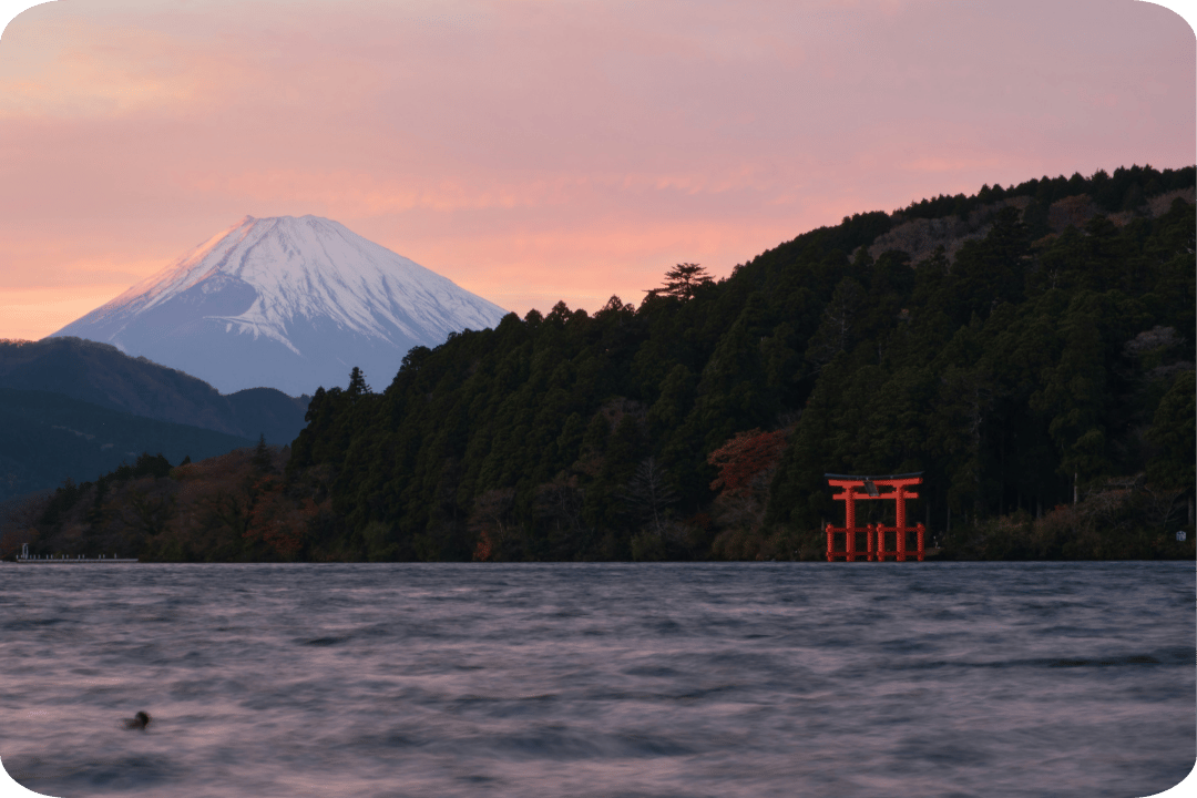 A view of Mount Fuji beyond a foreground of water, a red temple in front of the silhouette of dark trees and cliffs. 