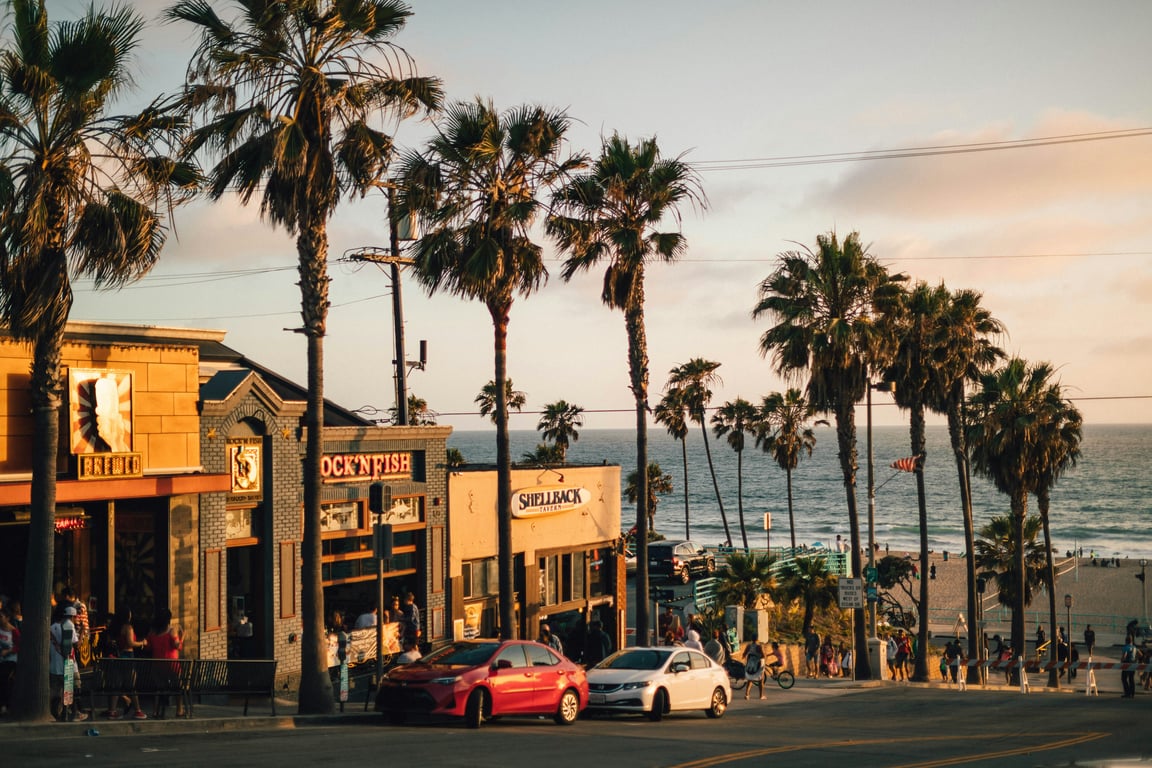 A road going downhill, palm trees are standing along its length, two cars are parked in the middle - one red, one white - people are scattered all over. At the bottom of the road is a beach. Along the road there are stores.