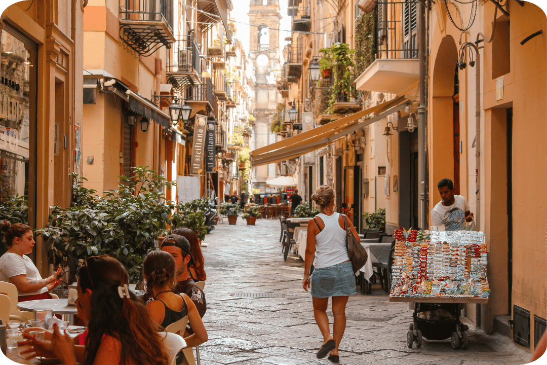 Some people sitting at tables outside a restaurant in a side road filled with potted plants. There are buildings on either side. A woman is walking away from the camera, a street seller is wheeling his cart down.
