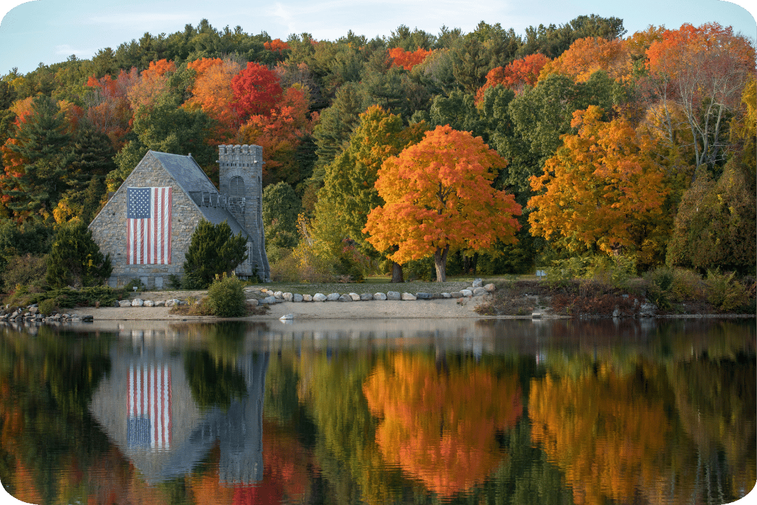 A small building by a lake with the American flag on it. It's surrounded by a tree-filled woodland, the trees are full and range from autumnal tones to greens.