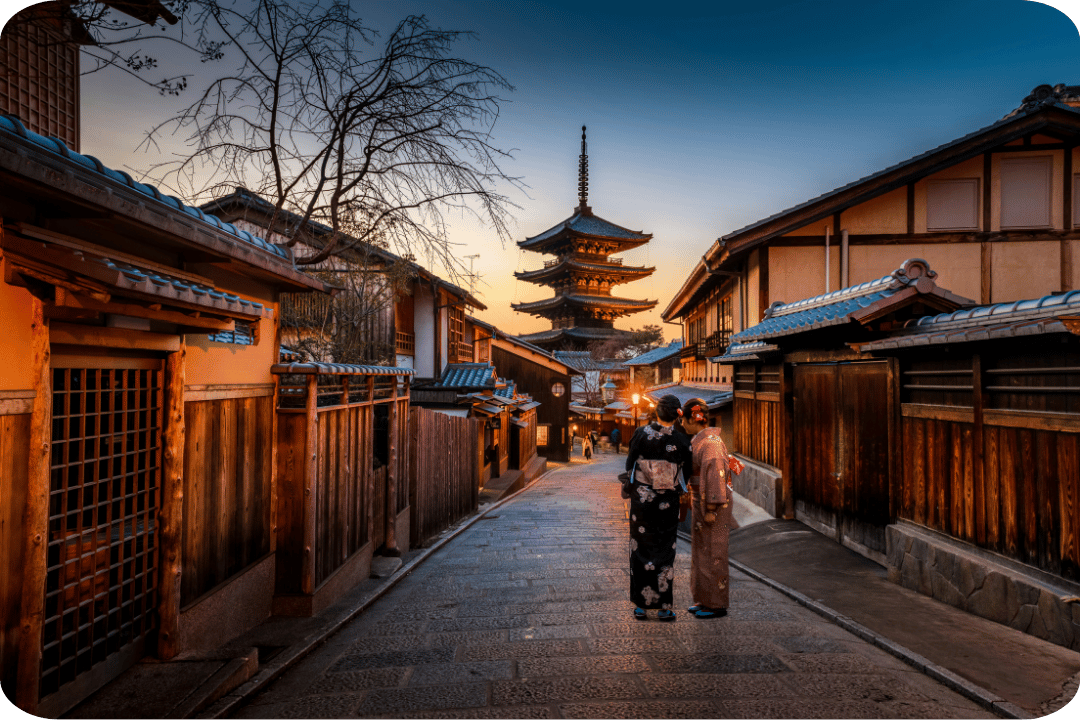 Two women photographed from behind leaning in to each other and talking on a street leading to a temple.