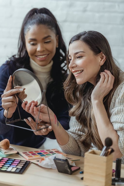 Cheerful young multiracial female friends with long wavy hair smiling and looking in round cosmetic mirror after applying makeup