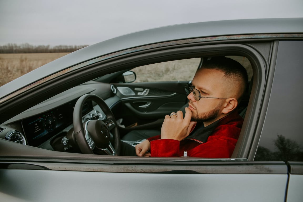 Vue latérale d'un jeune homme barbu et réfléchi portant des lunettes, assis sur le siège conducteur d'une voiture