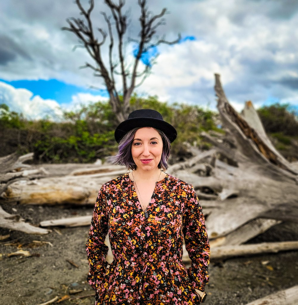 A close up shot of Elizabeth Jenkins on a beach along the Puget Sound. She is wearing a floral blouse and wearing a black hat. In the background, you can see some flotsam that has washed up upon the shore.