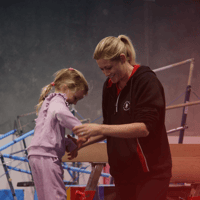Gymnast walking across beam, with coach helping them balance.