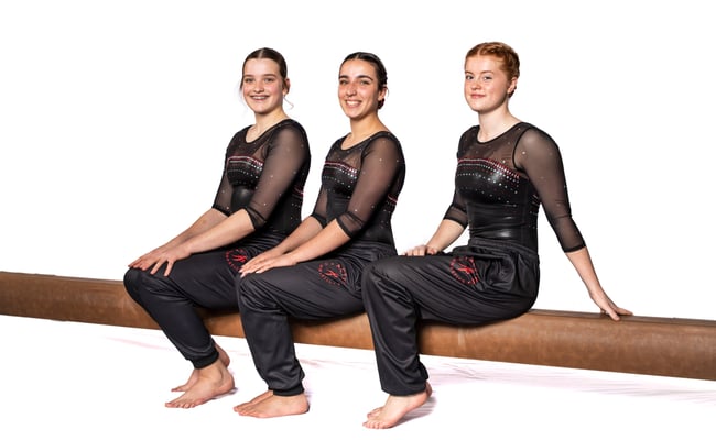 Three teenage gymnasts sitting on beam smiling at camera