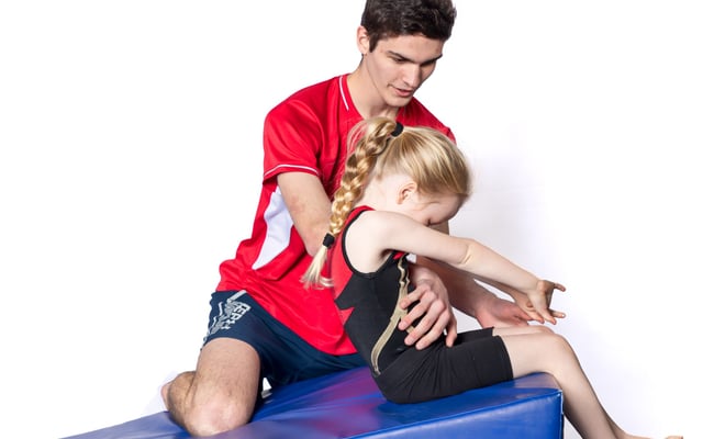 Gymnast starting a backward roll on a gymnastics wedge, with a coach helping to spot her.