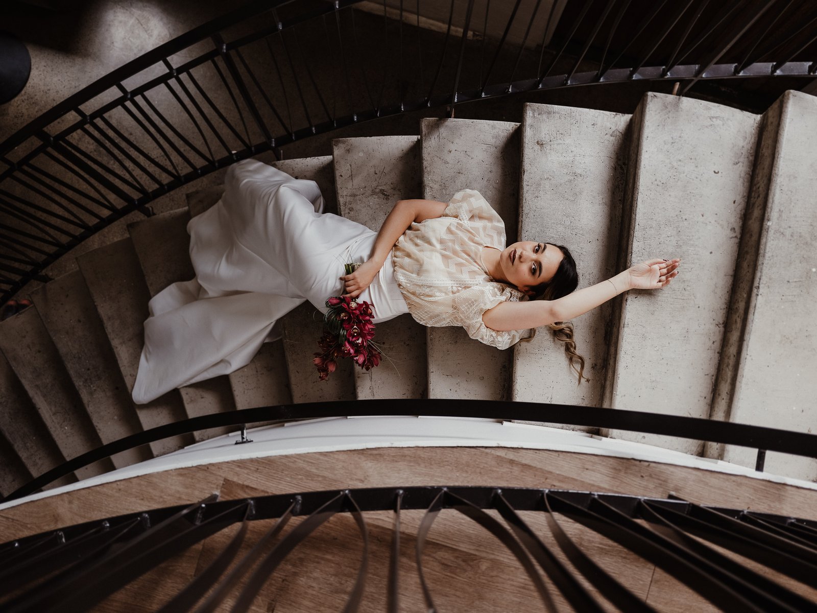 Bride in a white gown lying across the steps of a curved concrete staircase with a red bouquet.