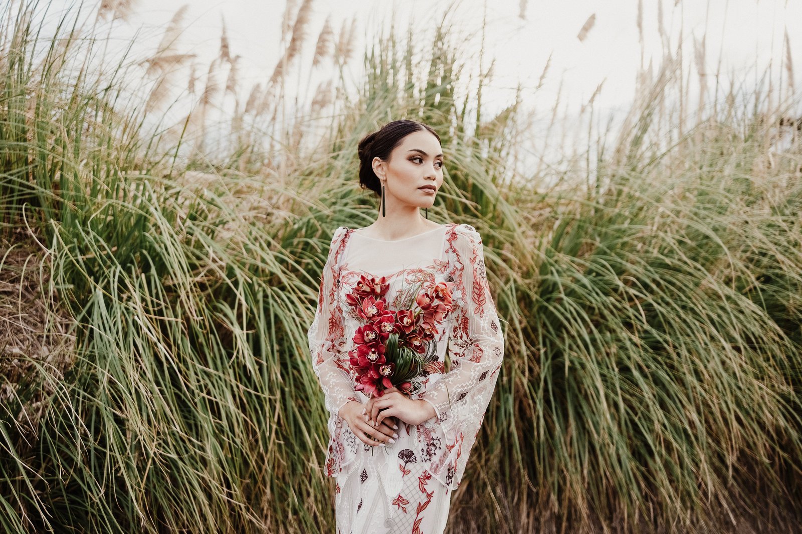 Bride holding a deep red bouquet while standing among toetoe in the red and white Huia gown.
