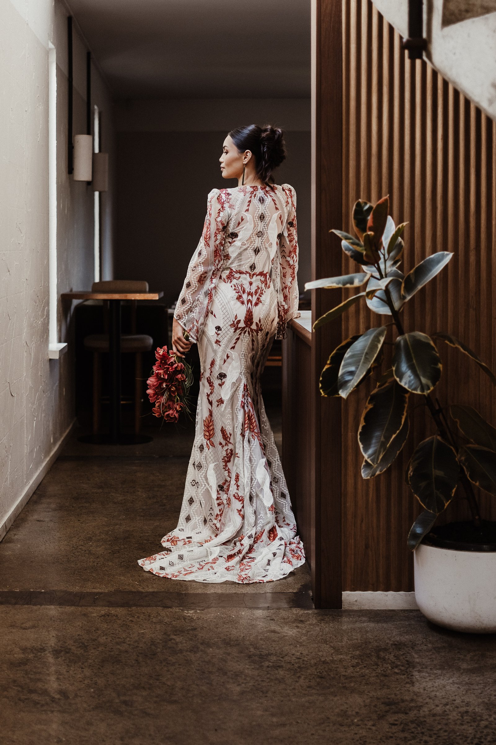 Bride in the red and white Huia gown standing in a timber-lined interior while holding a deep red bouquet.