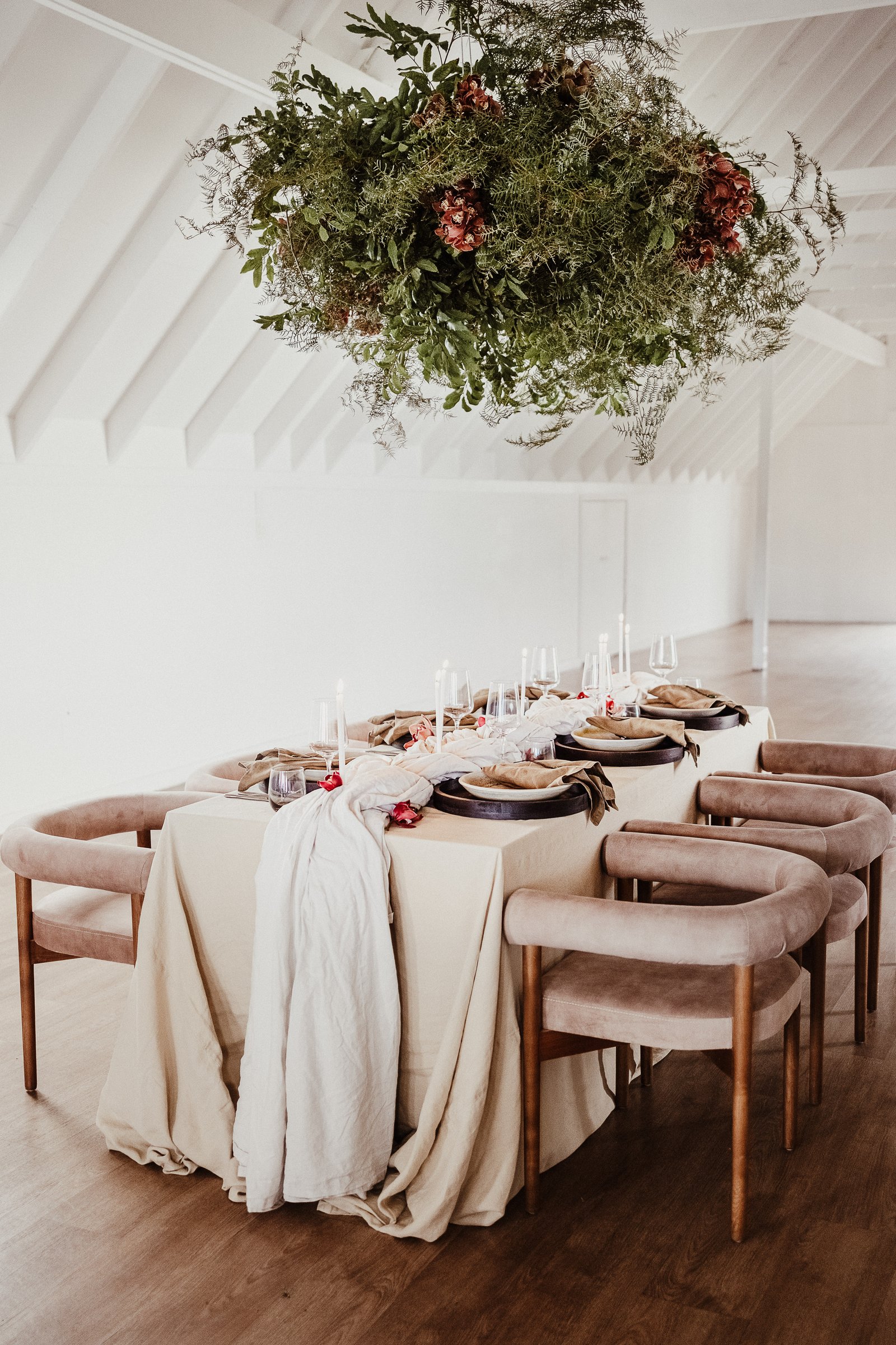 Wedding table set beneath a suspended installation of greenery and red flowers in a bright white venue.
