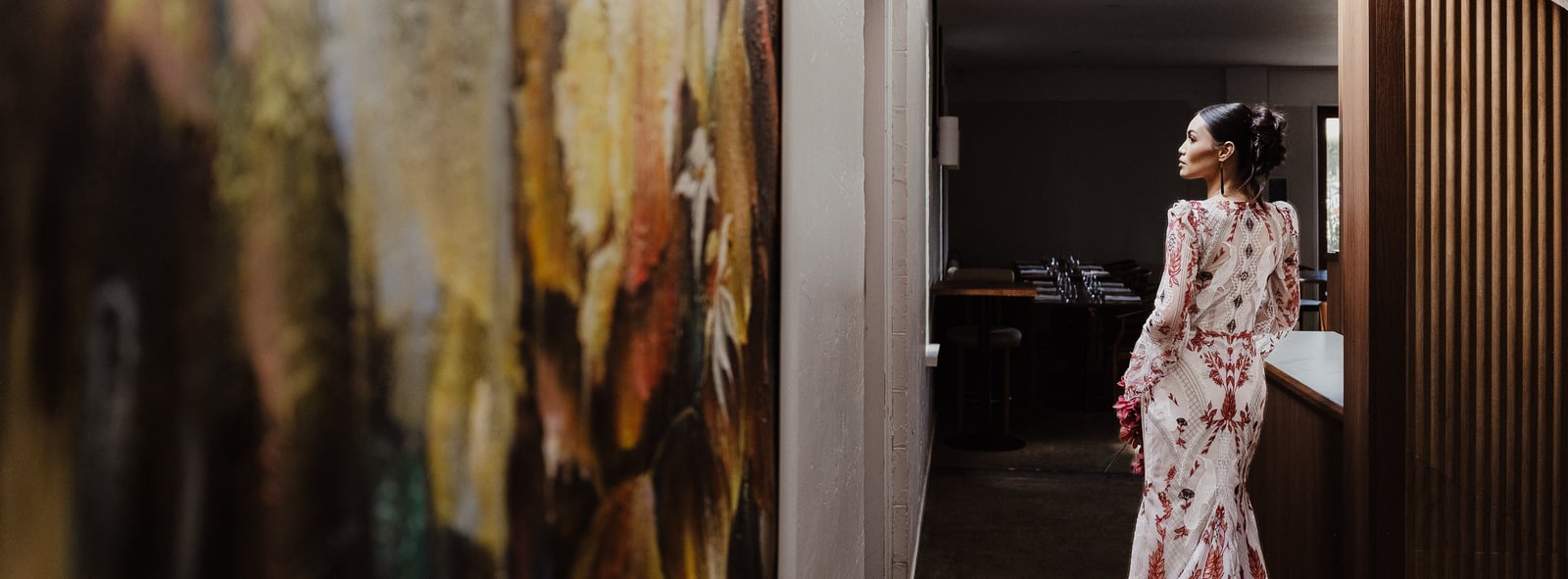 Bride in the Huia gown walking through a dark doorway beside a large artwork and timber wall.