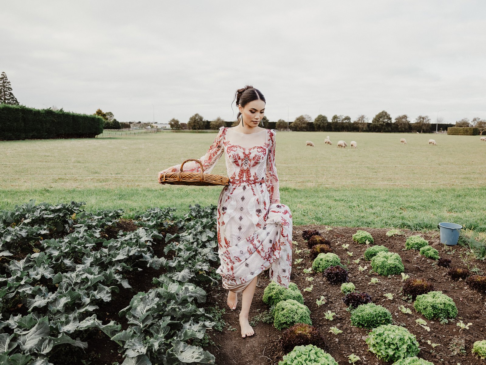 Bride in the Huia gown carrying a woven basket through a vegetable garden, with sheep in the field behind her.